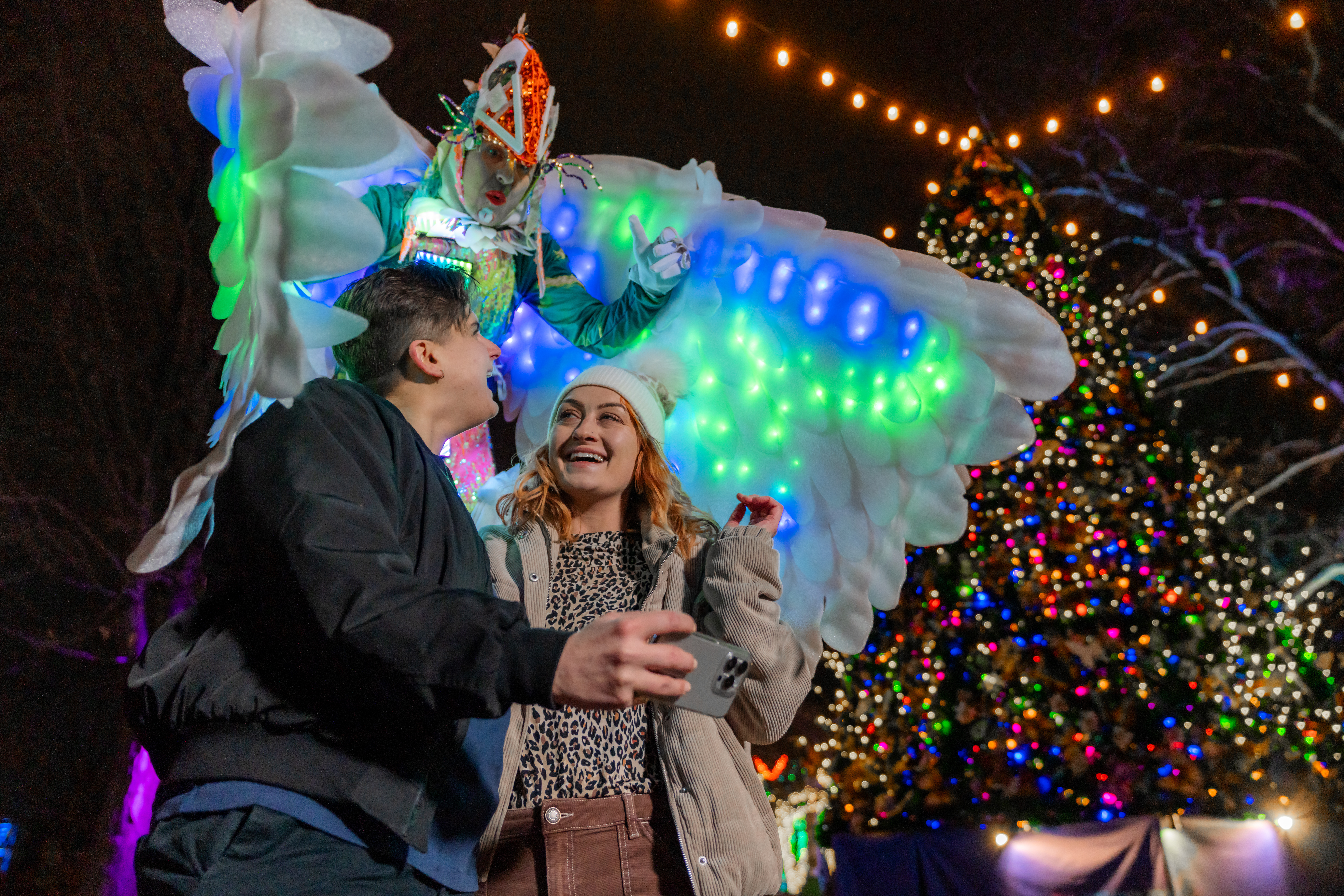 A couple takes a selfie next to a glowing tree with a light performer dressed as a bird at LumiNature, Philadelphia Zoo's holiday light show.